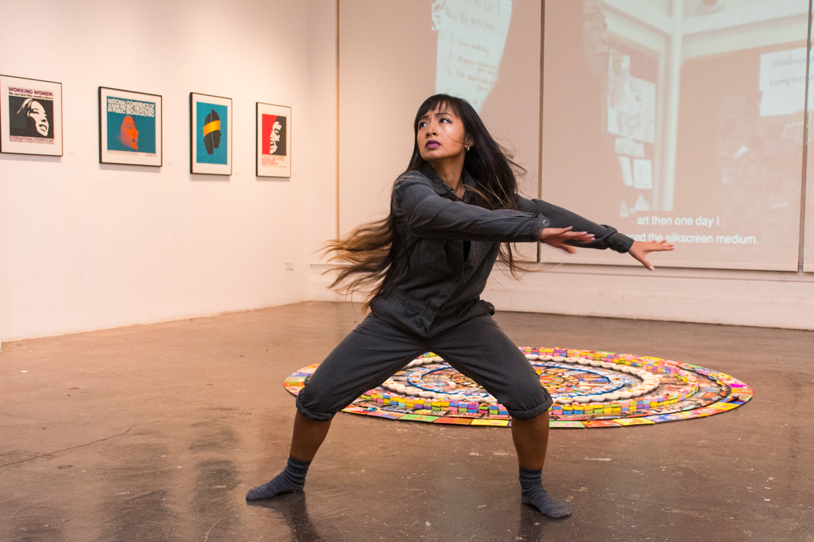 Female dancer stands with her arms outstretched  in front of a mandala. 