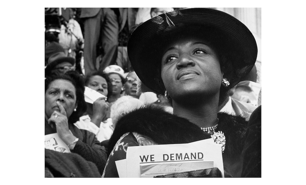 Image of a Black woman in a crowd wearing a black hat holding a stack of papers which read We Demand.