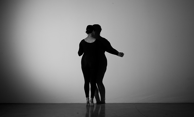 Image of a woman in a black unitard tracing her shadow against a wall.