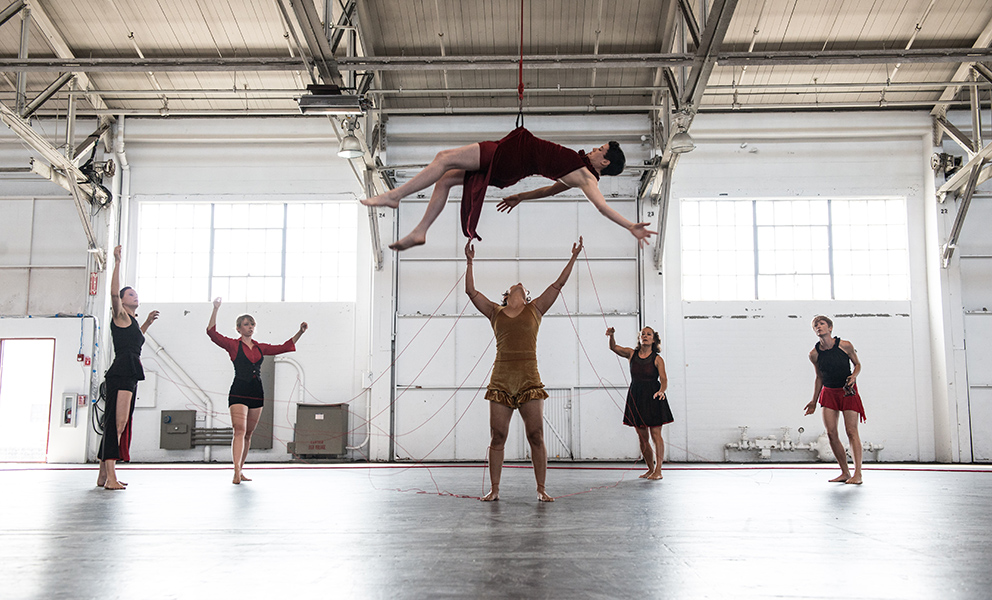 A dancer in a reclining position is suspended from the ceiling, while four other dancer watch from below