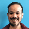A Latinx man with goatee and smiling, facing forward with a blue background.