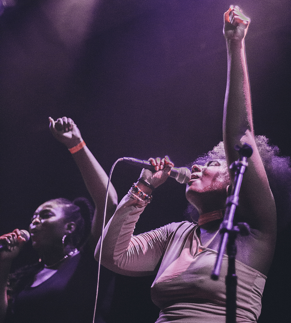Two Black women on stage holding mics to their mouths and singing with their fists up in the air. 