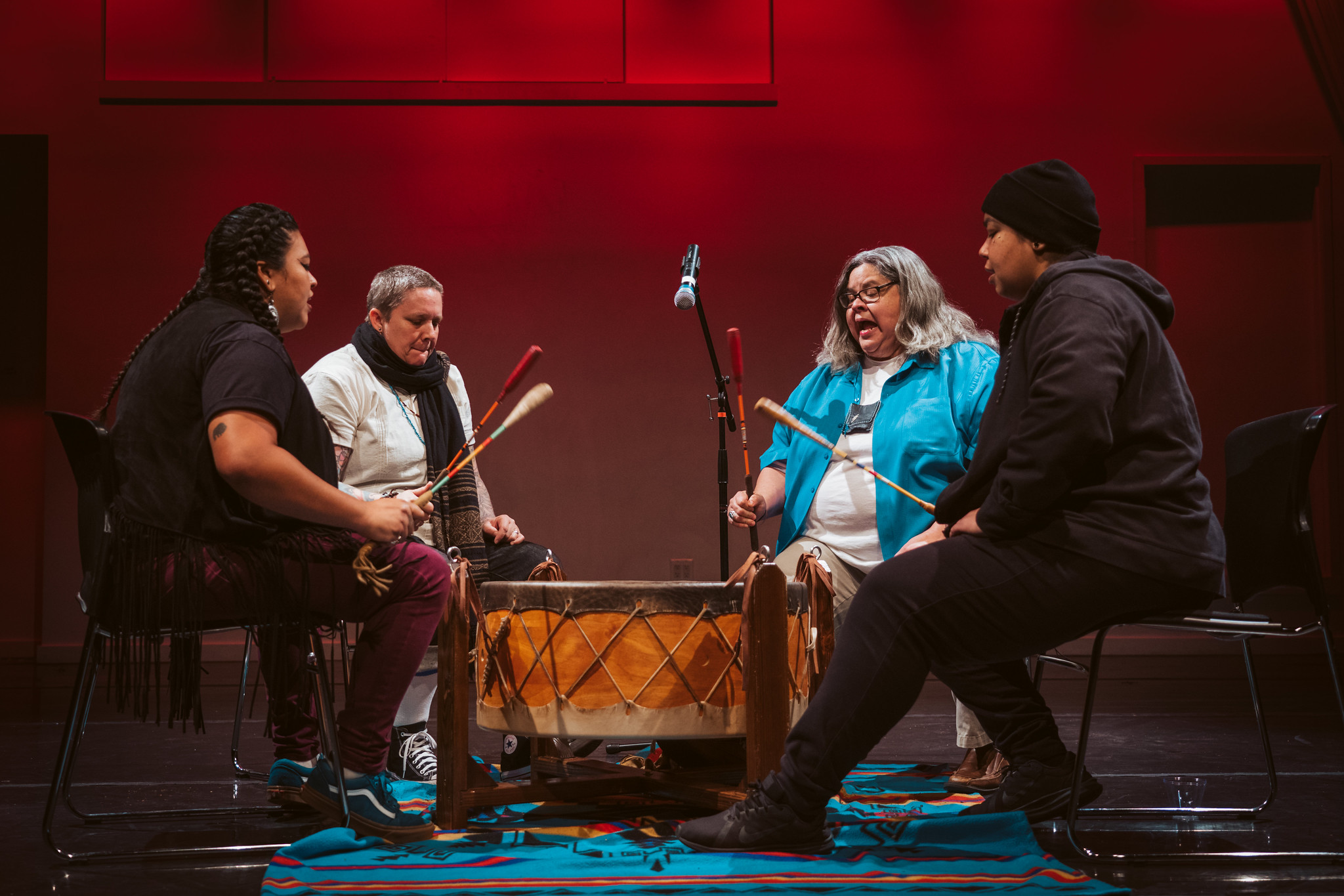 Four people circled around a large drum on a stage. Three of them have their mouths open to sing.