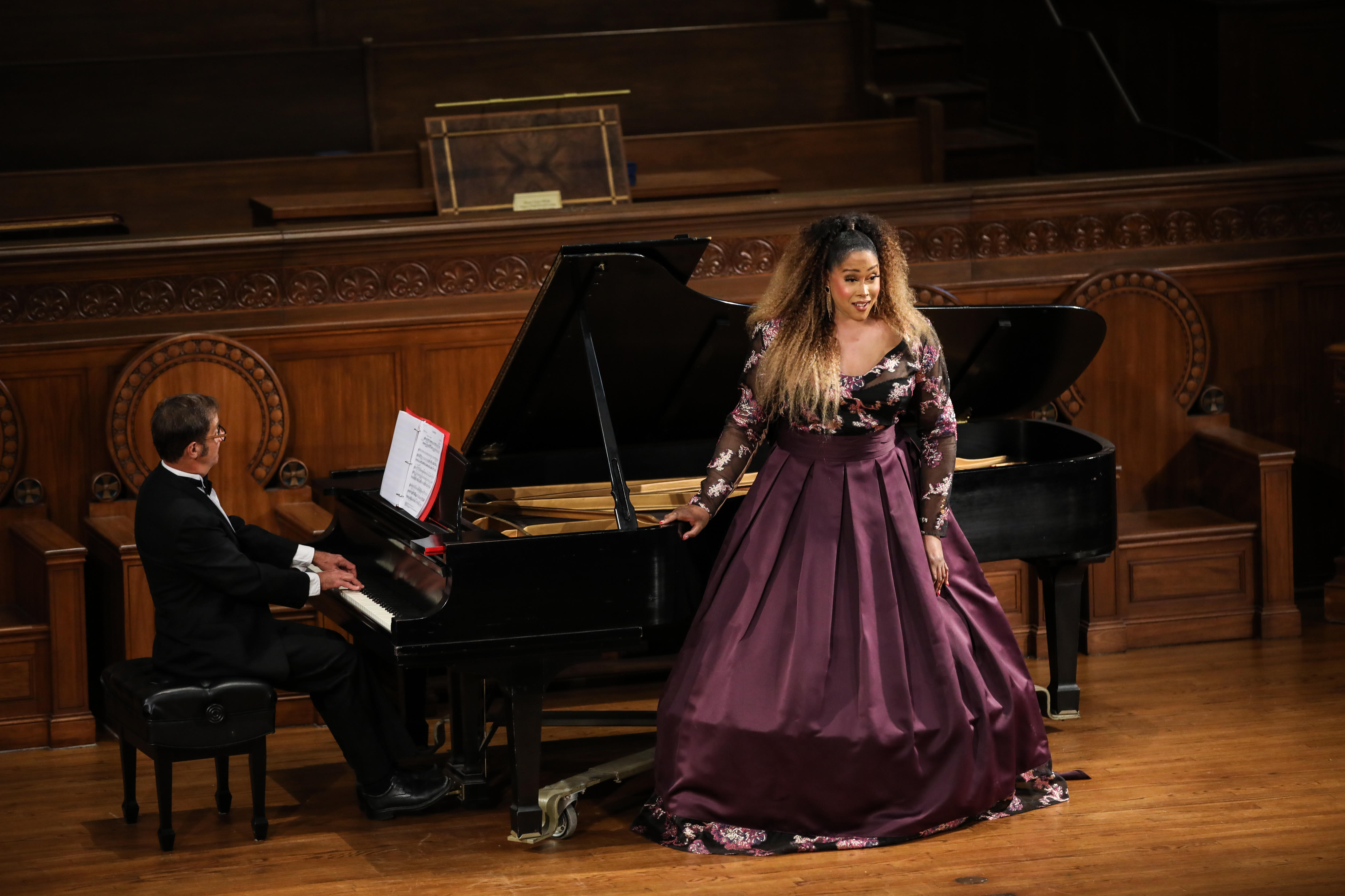Black woman opera singer with long blond and black hair in a floor length long-sleeved hooped burgundy dress standing in front of a grand piano on stage