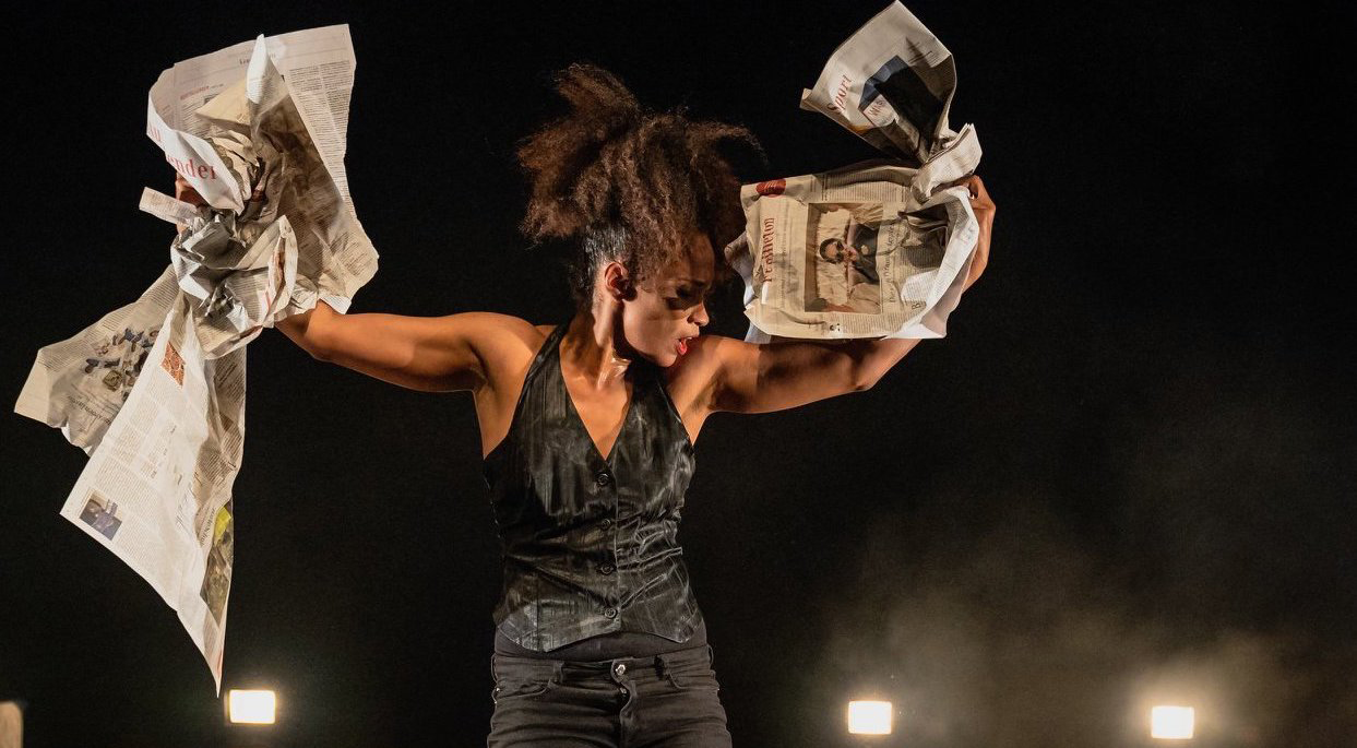 Black woman with high ponytail dressed in black jeans and black vest has hands raised above her head with crumpled newspapers in each and stands on a blacked out stage lit up by bright flood lights on the ground.