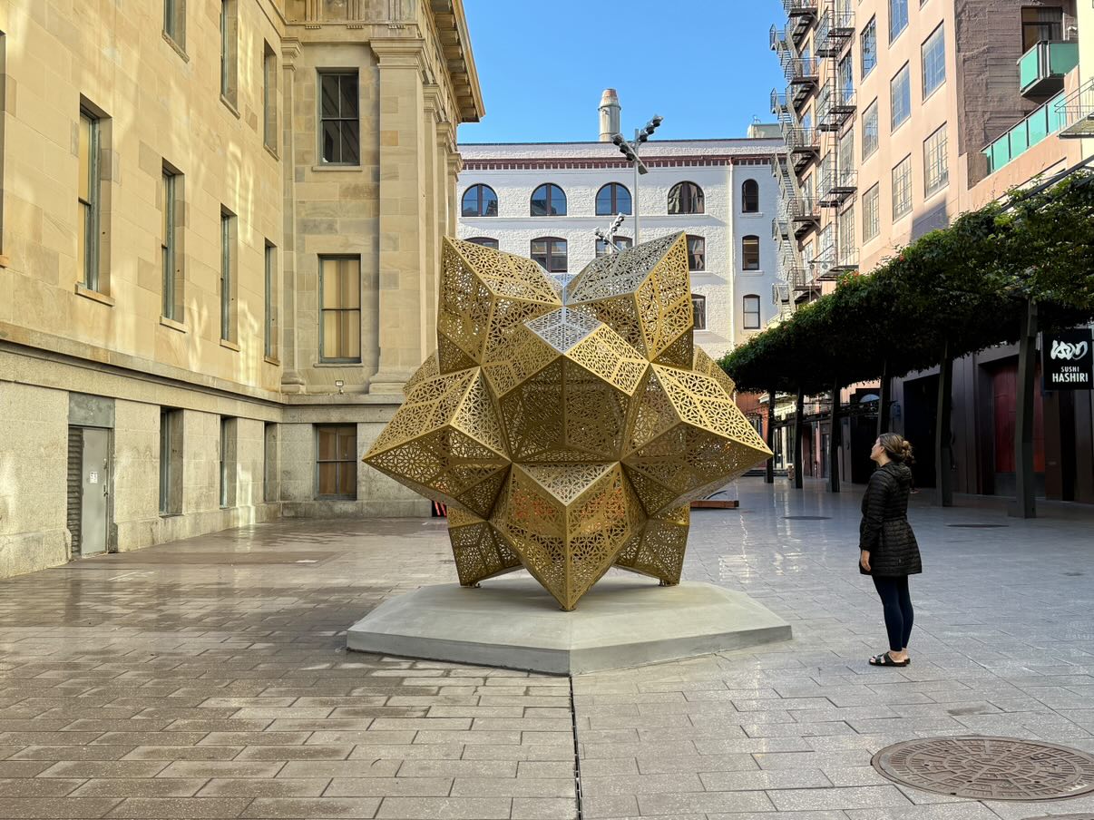 Image of Aurum sculpture in Mint Plaza in the daytime with a person standing next to it.