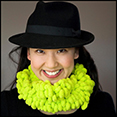 An Asian woman wearing a black fedora hat, facing forward and smiling. She is also wearing a bright lime green textured scarf. 
