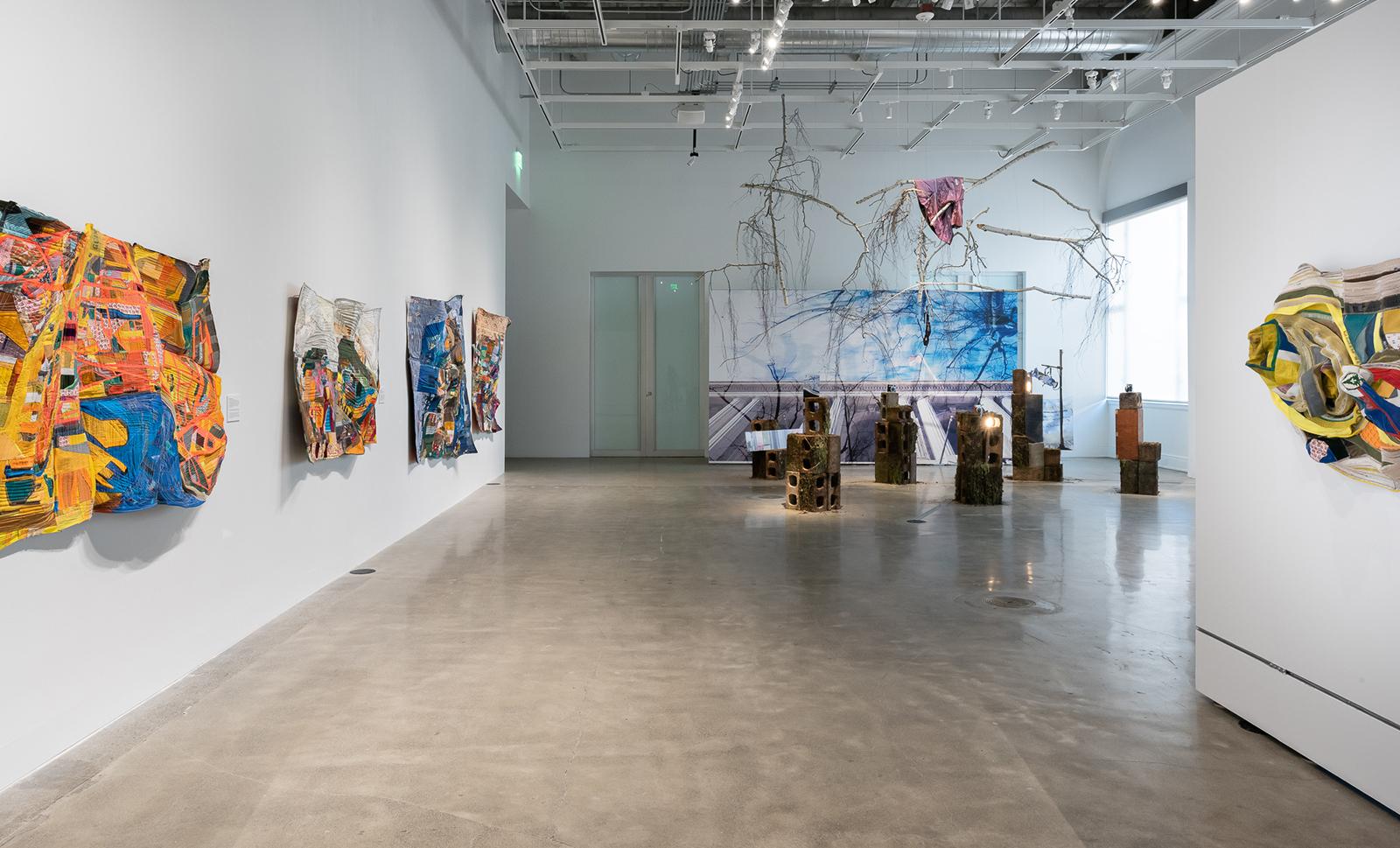 A view of Taking Place from the entrance of the gallery. On the left wall, are textile works by Mansur Nurullah. At the back is an installation by Hannah Waiters.